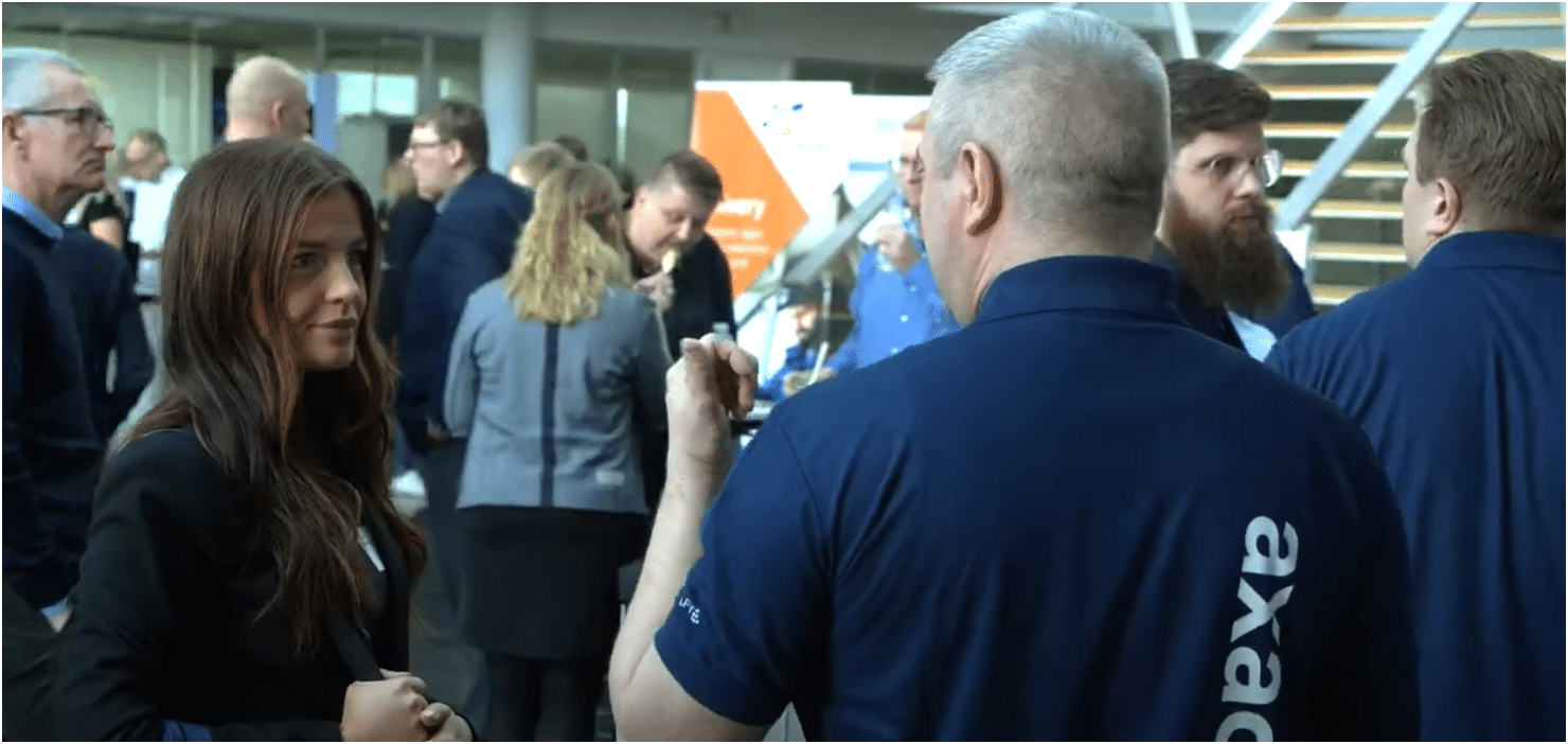 Video still fra Supply Chain experience day. Mørkhåret kvinde i front taler med lyshåret mand med ryggen til kamera. Axacon trykt på trøjen.